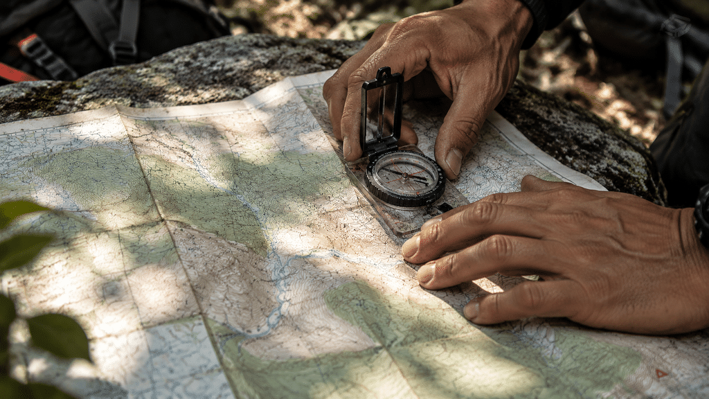 Tangan pendaki menggunakan peta topografi kertas dan kompas prisma manual untuk navigasi darat.